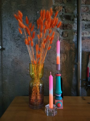 Bunte Kerzen in schicken Kerzenständern neben einer Glasvase mit orangfarbenen Trockenblumen.